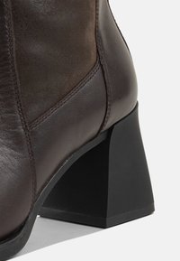 Brown leather ankle boot with a textured finish, featuring a tapered black heel and contrasting smooth side panels.