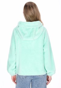 Világoszöld polár kapucnis pulóver puha tapintású anyagból, elülső zsebbel és állítható kapucnival. Laza szabású, enyhén ballon ujjakkal, világos farmerrel párosítva.