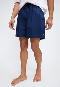 Άνδρας που φοράει ναυτικό μπλε μαγιό και λευκό μπλουζάκι, όρθιος ξυπόλυτος μπροστά σε απλό φωτεινό φόντο.