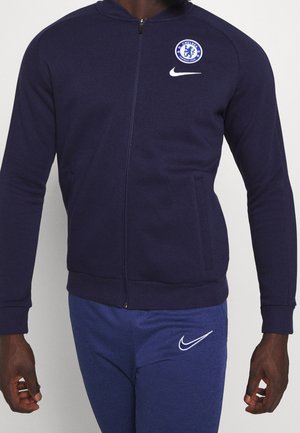 Mężczyzna ubrany w granatową kurtkę na zamek klubu piłkarskiego Chelsea oraz pasujące spodnie Nike z widocznymi logo na klatce piersiowej i udzie.