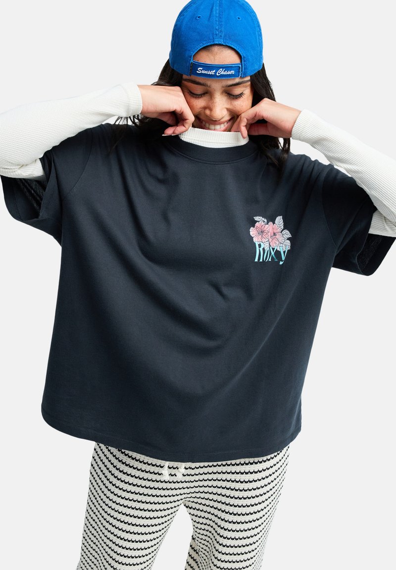 T-shirt en coton noir à manches courtes, avec un motif floral. Associé à un pantalon blanc et noir à motifs. Casquette bleue portée à l'envers.