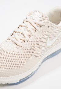 Gros plan sur une chaussure de sport beige Nike Zoom Air avec un dessus en maille, un logo swoosh blanc et des détails de semelle bleu translucide.