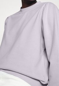 Lila sweatshirt med rund hals och puffärmar, gjord av mjukt tyg. Passformen är avslappnad, ihop med vita byxor.