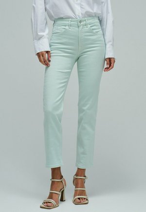 Mujer con vaqueros ajustados de color verde claro, camisa blanca abotonada y sandalias de tacón beige de pie frente a un fondo liso.