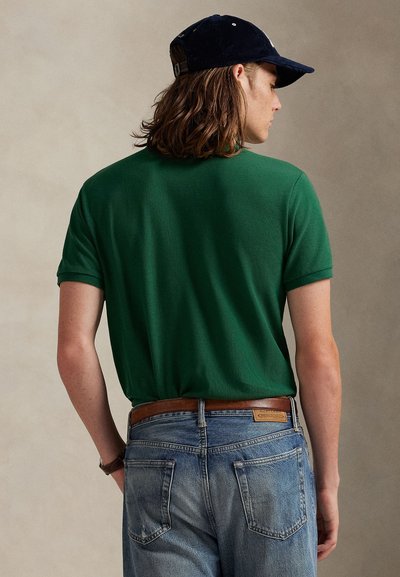 Muškarac s dugom kosom nosi crnu kapu, zelenu majicu kratkih rukava, smeđi pojas i plave traperice, snimljen odostraga na jednobojnoj pozadini.