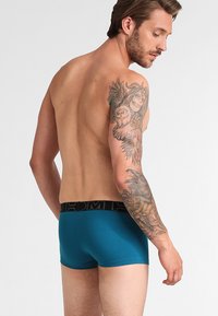 Tyrkysové boxerské trenýrky s černým brandingovým pasem, s hladkým povrchem. Model má tetování na viditelné pravé paži.