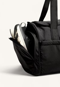 Zwarte duffeltas met een zijvak met rits waarin deels een zwarte sneaker zichtbaar is, gemaakt van stof met stevige stiksels.