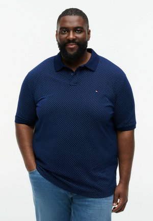 Smilende mand med skæg iført en mørkeblå mønstret polotrøje og lyseblå jeans, stående med den ene hånd i lommen.