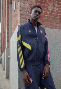 Mørkeblå træningsdragt med lynlåsjakke med gule og lyserøde detaljer, foret ærmer og broderet lyserødt logo på bukserne. Letvægtsmateriale.