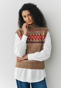 Hnědá pletená roláková vesta s červenými, bílými a modrými geometrickými vzory, přehodnocená přes bílou košili s objemovými rukávy a modrými džíny.