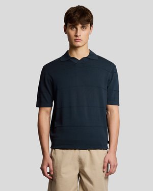 Ung mann iført en mørk marineblå kortermet poloskjorte og beige bukser, stående mot en ensfarget grå bakgrunn.