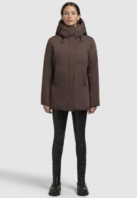 Brown hooded jacket with a straight cut, two front pockets, and drawstring features. Worn over black slim-fit leggings and boots.