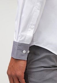 Camicia bianca con polsini grigi; presenta una chiusura a bottoni e una texture di tessuto liscia. I polsini hanno due bottoni e un dettaglio di cucitura in contrasto.