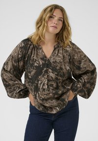 Femeie cu păr blond ondulat, purtând o bluză lejeră cu mâneci lungi, închisă cu nasturi, cu modele abstracte maronii și negre, și blugi de culoare albastru închis.