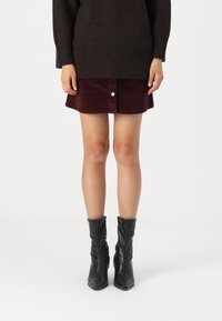 Bordeaux ribfluwelen rok met knoopsluiting aan de voorkant, gecombineerd met een donkere oversized trui en zwarte enkellaarsjes met een lichte hak.
