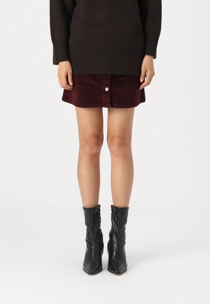 Bordeaux ribfluwelen rok met knoopsluiting aan de voorkant, gecombineerd met een donkere oversized trui en zwarte enkellaarsjes met een lichte hak.