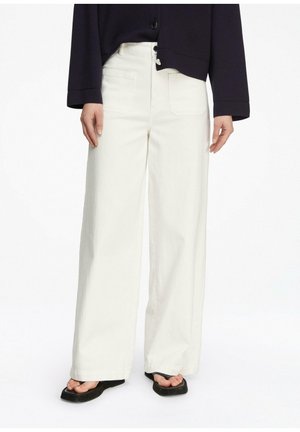 Osoba w białych spodniach o szerokich nogawkach, czarnej kurtce i czarnych sandałach na kwadratowym nosku, stojąca na białym tle.