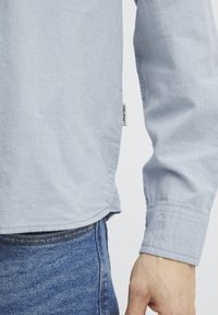 Lys blå langermet skjorte med teksturert stoff, knappbar mansjett og en synlig merkeetikett på siden, kombinert med tettsittende denimjeans.