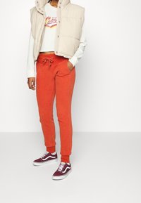 Béžový péřový vest s bílou zkrácenou mikinou s barevným potiskem, doplněný červenými tepláky a vínovými nízkými teniskami.