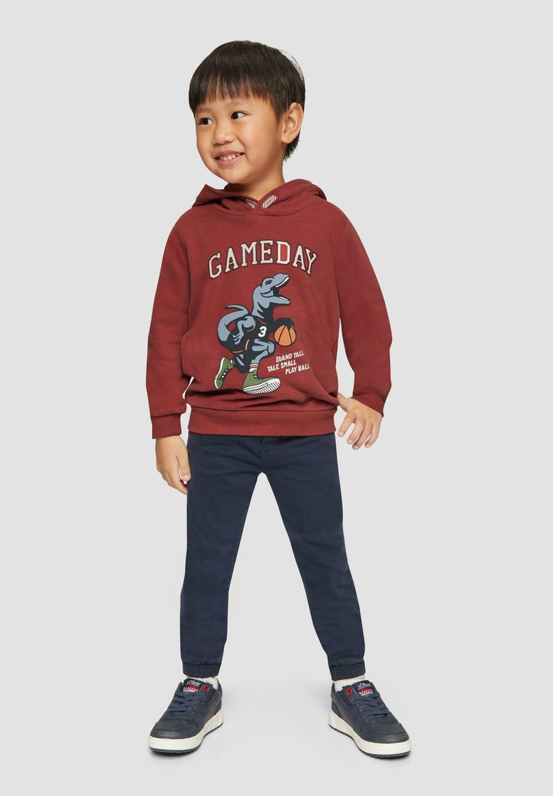 Czerwona bluza z kapturem z grafiką dinozaura i napisem "GAMEDAY", w połączeniu z ciemnoniebieskimi spodniami i niebieskimi trampkami z białymi podeszwami.