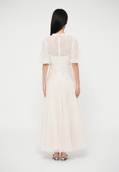 Μακρύ, ανοιχτό μπεζ φόρεμα με διάφανα φουσκωτά μανίκια και υφασμάτινες λεπτομέρειες με υφή, με εφαρμοστή μέση και ροή φούστας. Στο πίσω μέρος διαθέτει κούμπωμα με κουμπιά.