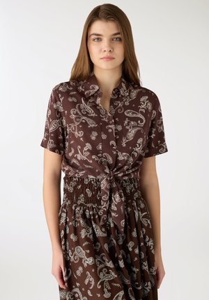 Giovane donna che indossa una camicia marrone a maniche corte con motivo paisley, annodata in vita, e una gonna vita smock abbinata, su uno sfondo semplice.