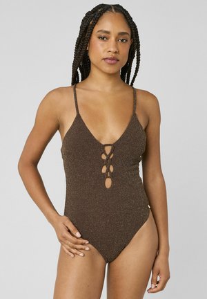 Femme aux longues tresses portant un maillot de bain marron texturé avec un devant lacé et de fines bretelles, debout devant un fond gris uni.
