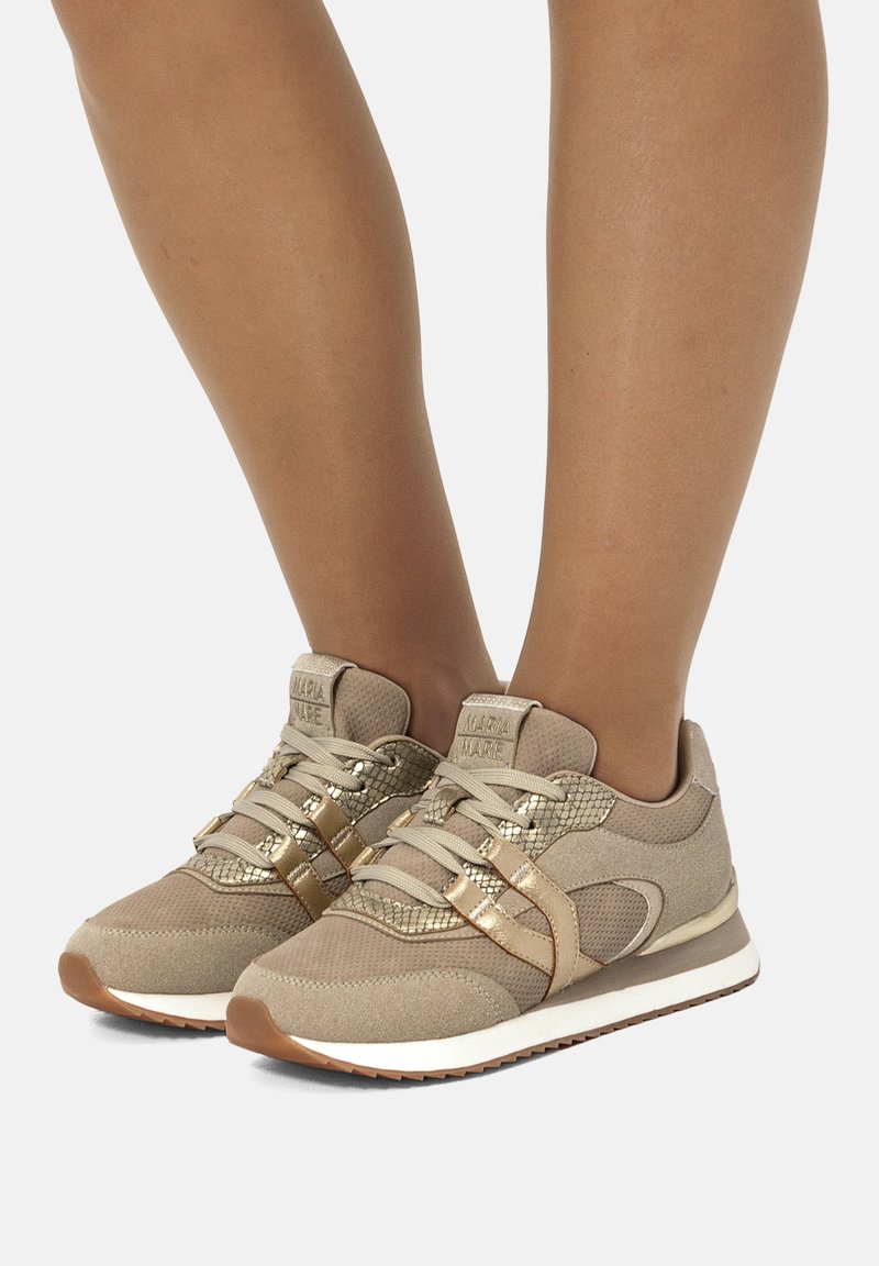 Beige Sportschuhe zeichnen sich durch eine Mischung aus strukturierten Stoffen, goldenen Akzenten und einer weißen Sohle aus. Das Design beinhaltet Schnürsenkel und gemusterte Details.