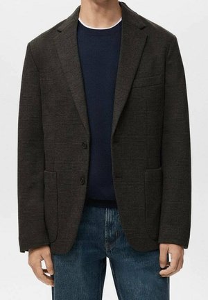 Mann trägt dunkel karierten Blazer über marineblauem Pullover und blauen Jeans, steht mit entspannten Händen an den Seiten.