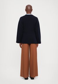 Person som står med ryggen mot kameran, klädd i en mörk marinblå tröja, vida bruna byxor och svarta skor framför en neutral bakgrund.