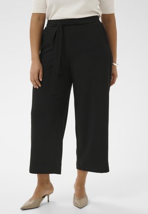 Femme portant un pantalon noir large et court avec ceinture nouée à la taille et escarpins beige à bout pointu et petit talon, debout devant un fond uni.
