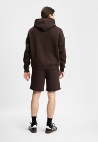 Brun hettegenser og matchende shorts laget av mykt stoff, med en avslappet passform og ribbestrikkede mansjetter, kombinert med svarte og hvite sko.