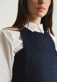 Robe tablier en denim bleu superposée sur une chemise blanche à col. Présente un corsage structuré et une texture lisse avec des détails de couture minimaux.