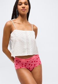 Γυναίκα με μακριά κοτσίδα, που φοράει λευκό αμάνικο τοπ με διάτρητο σχέδιο και ροζ εσώρουχο με σχέδιο καρδιές στη μέση με το λογότυπο "PIECES".