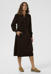 Braunes Cordkleid mit Knopfleiste, langen Ärmeln, Seitentaschen und einem Saum unterhalb des Knies. Glatte Textur mit weichem Finish.
