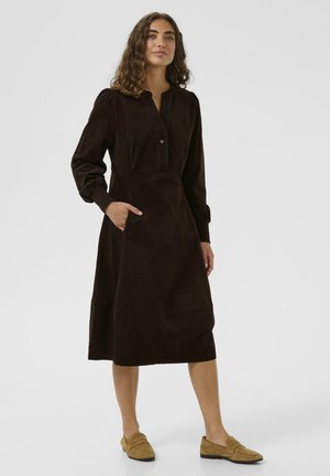 Robe en velours côtelé marron avec une patte de boutonnage, des manches longues, des poches latérales et une longueur sous le genou. Texture lisse avec une finition douce.
