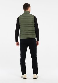 Grüne Steppweste mit hohem Kragen, ausgestattet mit einem gesteppten Design und glatter Textur. Getragen über einem schwarzen Langarmshirt und kombiniert mit schwarzen Hosen.