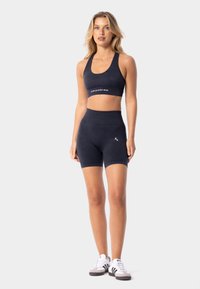 Jūras spēku sporta krūšturis un augsto jostasvietu šortus komplekts, kas izgatavots no mīkstas auduma, ar logotipu uz jostas un īsu apakšmalu. Tērps kombinēts ar baltām kedām.