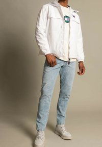 Giacca leggera bianca con polsini elasticizzati, abbinata a una t-shirt di colore chiaro e jeans blu sbiaditi. Il modello indossa scarpe alte grigie.