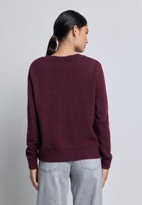 Femme avec une queue de cheval noire portant un pull long à manches marron et un jean gris clair, de dos contre un fond clair uniforme.