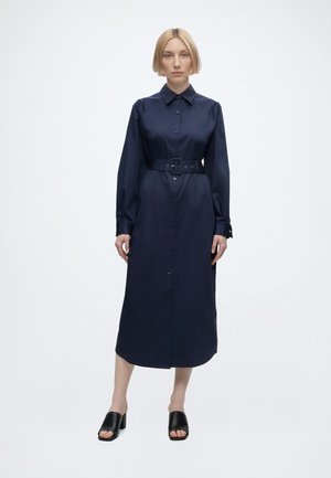 Femme aux cheveux blonds coiffée d’un carré, portant une robe-chemise bleu marine à manches longues ceinturée et des mules noires à talons ouverts, debout devant un fond uni.