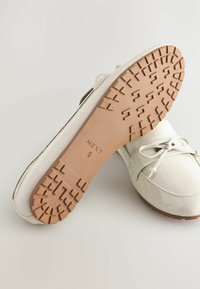 Scarpa da barca in pelle sintetica bianca con punta rotonda, lacci e suola in gomma testurizzata con motivi a rilievo geometrici.