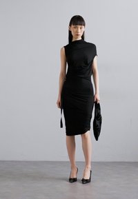 Vestido preto justo com um design drapeado e decote assimétrico. Combinado com uma clutch preta texturizada e sapatos de salto alto.