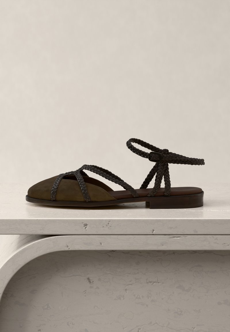 Καφέ επίπεδο σανδάλι με πλεκτούς ιμάντες και αγκράφα, κλειστή μύτη, εκτεθειμένο σε φωτεινή υφασμάτινη επιφάνεια.