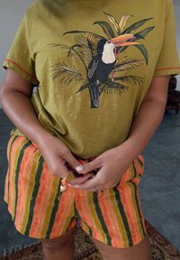 Olive green shirt with a toucan illustration, paired with orange and green striped shorts. Features a drawstring waist and lightweight fabric.