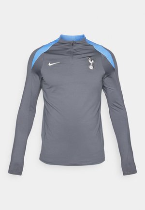 Grå langærmet sportstrøje med kvart lynlås, der har blå skulderaccenter og et hvidt Nike-logo samt et Spurs-emblem på brystet.