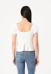 Blouse blanche en coton avec des manches bouffantes, dos smocké et ourlet peplum. Associée à un jean en denim bleu avec poches arrière et coutures.