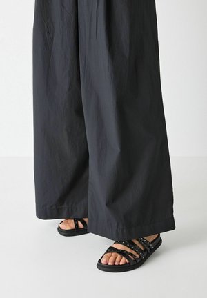 Füße in schwarzen flachen Sandalen mit mehreren Riemen und kleinen Nieten, kombiniert mit locker sitzenden schwarzen Hosen mit weitem Bein.