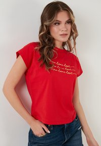 Rød bomulds t-shirt med korte ærmer, prydet med sætningen "no love lost" i guld. Casual pasform med rund halsudskæring.