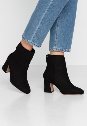Kvinde iført sorte ruskindsstøvler med blokhæl og lyseblå cropped jeans står på en hvid gulv.